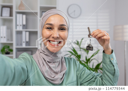 A cheerful woman in a hijab proudly showing a house key, symbolizing new home ownership and a fresh start in her cozy indoor setting. A cheerful woman in a hijab proudly showing a house key, symbolizing new home ownership and a fresh start in her cozy indoor setting. 114779344