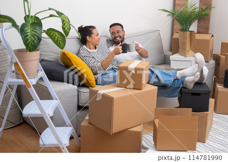 Unpacking and moving day become a celebration as a young couple cheers with coffee cups on the sofa, surrounded by boxes, making their new house their own 114781990