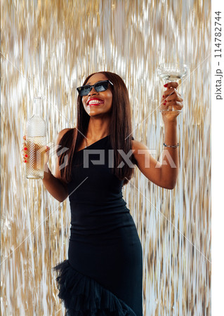 Young happy fashion African girl black woman in cocktail dress and sunglasses holding drink glass and wine bottle dancing at party with glitter background having fun at holiday, birthday celebration. Young happy fashion African girl black woman in cocktail dress and sunglasses holding drink glass and wine bottle dancing at party with glitter background having fun at holiday, birthday celebration. 114782744