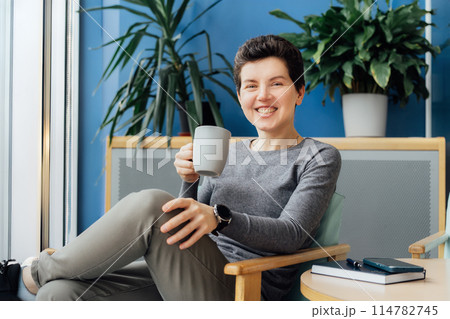 Smiling Neutral gender middle aged business woman in casual clothing relaxing with coffee cup on the armchair. Female professional enjoying coffee drink in office lounge. Take a break for recovery Smiling Neutral gender middle aged business woman in casual clothing relaxing with coffee cup on the armchair. Female professional enjoying coffee drink in office lounge. Take a break for recovery 114782745