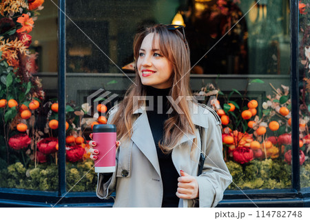Young smiling woman in trench coat enjoying drink in reusable coffee cup walking on street with shop windows decorated for the autumn holidays. Happy fall cozy mood, seasonal fashion style trend 114782748