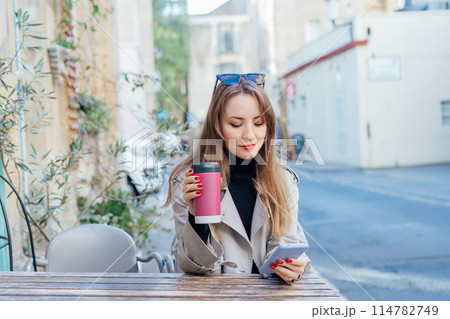 Elegant young woman using phone and drink coffee from reusable cup sitting outdoors. Texting resting in cafe terrace table after city walk. using internet in wifi zone, enjoying coffee and leisure. Elegant young woman using phone and drink coffee from reusable cup sitting outdoors. Texting resting in cafe terrace table after city walk. using internet in wifi zone, enjoying coffee and leisure. 114782749