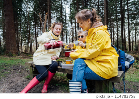 Halt for snack during hiking. Company hikers enjoying picnic on bench, drinking hot tea, eating sandwiches in forest. Friends relaxing and having snack picnic on nature background. Backpacker trip. Halt for snack during hiking. Company hikers enjoying picnic on bench, drinking hot tea, eating sandwiches in forest. Friends relaxing and having snack picnic on nature background. Backpacker trip. 114782758