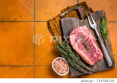Raw Wagyu striploin, club steak on wooden board with thyme and rosemary ready for cooking. Orange background. Top view. Copy space Raw Wagyu striploin, club steak on wooden board with thyme and rosemary ready for cooking. Orange background. Top view. Copy space 114788138