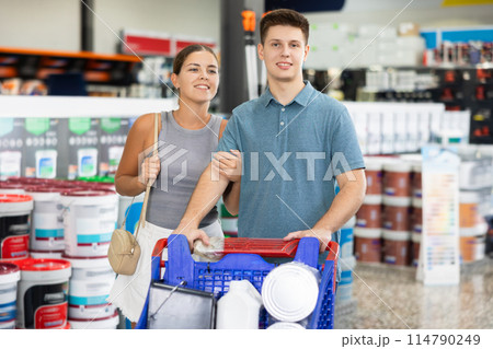 Smiling young woman and man with trolley looking for paints for house renovation and talking friendly while visiting construction hypermarket Smiling young woman and man with trolley looking for paints for house renovation and talking friendly while visiting construction hypermarket 114790249