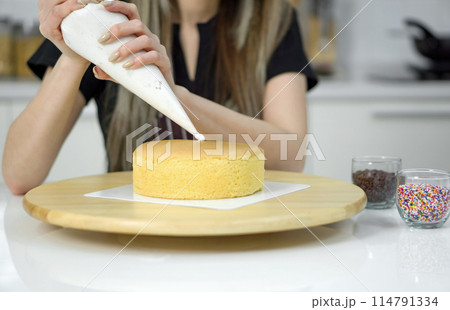 Closeup hand piping white cream on a sponge cake, creating a decorative pattern on the top of a round, light-colored cake that's placed on a wooden turntable or cake stand. Closeup hand piping white cream on a sponge cake, creating a decorative pattern on the top of a round, light-colored cake that's placed on a wooden turntable or cake stand. 114791334