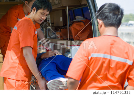 Group of paramedic or emergency medical technician (EMT) in an orange uniform places a neck and head accident victim on a bed in an ambulance. Urgent assistance during road accident. Group of paramedic or emergency medical technician (EMT) in an orange uniform places a neck and head accident victim on a bed in an ambulance. Urgent assistance during road accident. 114791335