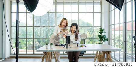 Two asian women creating content for social media. They are using a studio setup that include a smartphone mounted on a tripod, lighting equipment, and various makeup products arrayed on the table. Two asian women creating content for social media. They are using a studio setup that include a smartphone mounted on a tripod, lighting equipment, and various makeup products arrayed on the table. 114791916