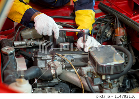 Closeup mechanic hand in work uniform, working on a car in an automotive shop or garage. He seem focused and engaged in diagnosing or repairing the vehicle's engine or other component. 114792116