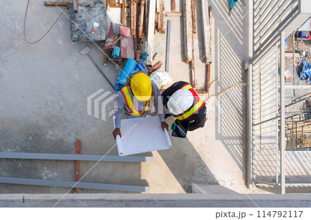 Two worker in hardhat and reflective vest are busy, looking at blueprint on a sunny day at a construction site. High angle view. 114792117