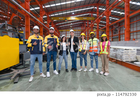 A team of worker stand together for a group photo inside a factory with machinery in the background. Everyone smiling and raise finger thumbs up. 114792330