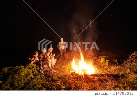 Young family drinking tea next to fire place. Cozy night in forest. Family of four warming near campfire. 114793013