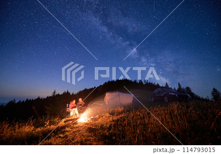 Night camping in the mountains under starry sky full of stars. Travelling couple having a rest at campfire, tourist tent and off-road vehicle in the evening on mountain lawn and stargazing together. 114793015