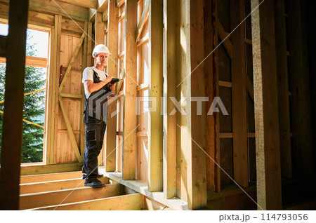 Carpenter constructing wooden framed house. Man worker working with screwdriver, wearing work overalls and helmet. Concept of modern eco-friendly construction. Carpenter constructing wooden framed house. Man worker working with screwdriver, wearing work overalls and helmet. Concept of modern eco-friendly construction. 114793056