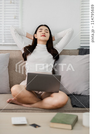 A woman is sitting on a couch with a laptop on her lap. She is wearing a white shirt and she is relaxed 114795689