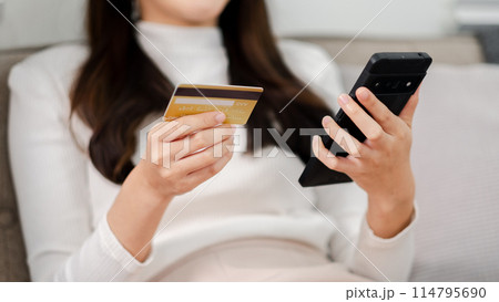 A woman is holding a credit card and a cell phone. She is sitting on a couch. Concept of modern life and convenience, as the woman is able to make a purchase using her credit card A woman is holding a credit card and a cell phone. She is sitting on a couch. Concept of modern life and convenience, as the woman is able to make a purchase using her credit card 114795690