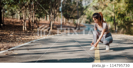 A woman is kneeling on the road, tying her shoe. The scene is set in a park with trees in the background A woman is kneeling on the road, tying her shoe. The scene is set in a park with trees in the background 114795730