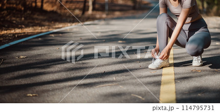 A woman is kneeling on the road, putting on her shoes. Concept of determination and focus as the woman prepares for her run 114795731