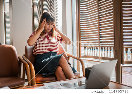 A woman sits in a chair with her head in her hands. She is wearing a striped shirt and blue jeans. A laptop is on a table in front of her A woman sits in a chair with her head in her hands. She is wearing a striped shirt and blue jeans. A laptop is on a table in front of her 114795752