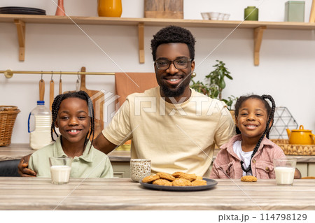 Happy African father and daughter eating cookies and drinking milk kitchen Happy African father and daughter eating cookies and drinking milk kitchen 114798129