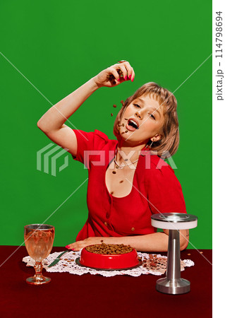 Quirky Meal Choice. Young woman in red dress eating from bowl of pet food against green background. Humorous dining Quirky Meal Choice. Young woman in red dress eating from bowl of pet food against green background. Humorous dining 114798694
