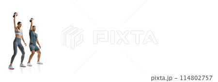 Banner. Man and woman dressed sportwear in mid-exercise, each holding kettlebell aloft with one arm against white studio background. Banner. Man and woman dressed sportwear in mid-exercise, each holding kettlebell aloft with one arm against white studio background. 114802757