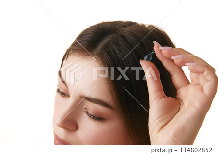 Close up of young brunette woman's head with silky, straight hair holding capsule with oil against white background. Close up of young brunette woman's head with silky, straight hair holding capsule with oil against white background. 114802852