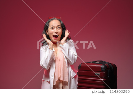Happy and excited senior woman with suitcase over red background. Summer, travel and vacation concept 114803290