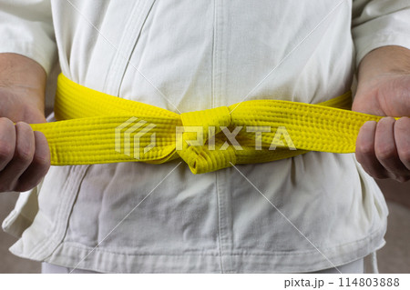 Male hands tighten yellow belt in Kyokushin karate, Asian types of martial arts with belts, the sixth kyu puts belt on karate kimono Male hands tighten yellow belt in Kyokushin karate, Asian types of martial arts with belts, the sixth kyu puts belt on karate kimono 114803888