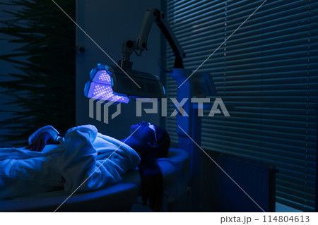 Remote view of female patient undergoes blue LED light therapy for skin rejuvenation at modern aesthetic clinic. Using phototherapy device with blue light wavelength for acne treatments. Remote view of female patient undergoes blue LED light therapy for skin rejuvenation at modern aesthetic clinic. Using phototherapy device with blue light wavelength for acne treatments. 114804613