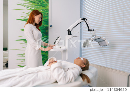 Professional female beautician specialist connecting and adjusting device for photo peeling at modern aesthetic clinic. Woman having LED light facial photodynamic therapy treatment in beauty salon. Professional female beautician specialist connecting and adjusting device for photo peeling at modern aesthetic clinic. Woman having LED light facial photodynamic therapy treatment in beauty salon. 114804620