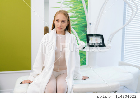 Portrait of pretty young woman patient in white bath robe sitting on medical couch in modern cosmetology clinic while waiting beautician specialist. Concept of cosmetology, spa, beauty treatment 114804622