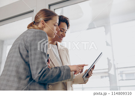 Smiling female business people standing in modern coworking and using digital tablet together Smiling female business people standing in modern coworking and using digital tablet together 114809310