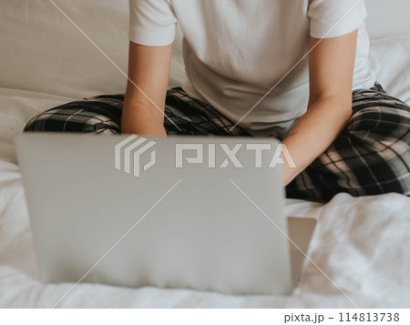 Uncertain woman with bent legs working at laptop in pajamas on bed in bedroom 114813738