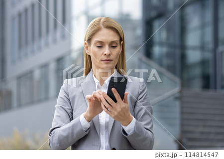 Focused businesswoman in gray suit browsing her smartphone against a modern office building backdrop. Captures a moment of technology use in a corporate environment. 114814547