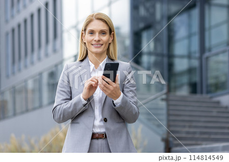 A confident professional woman in a business suit uses her smartphone while standing outside a modern office building. She appears content and focused. A confident professional woman in a business suit uses her smartphone while standing outside a modern office building. She appears content and focused. 114814549