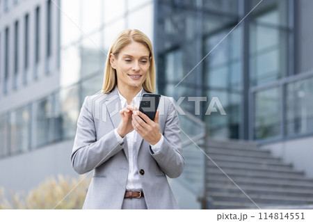 A professional woman in a gray suit smiles as she uses a smartphone outside a modern office building. Emphasizing connectivity and business efficiency. 114814551