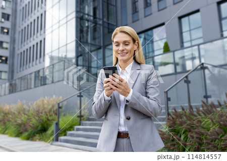 A businesswoman in a gray suit uses her smartphone outside modern office buildings, reflecting a blend of professionalism and technology in a corporate environment. 114814557
