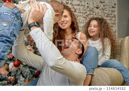 Christmas Family. Happiness. Portrait of dad, mom and children of different ages are sitting on the sofa at home near the Christmas tree and fireplace, everyone is smiling. New Year's happiness Christmas Family. Happiness. Portrait of dad, mom and children of different ages are sitting on the sofa at home near the Christmas tree and fireplace, everyone is smiling. New Year's happiness 114814582