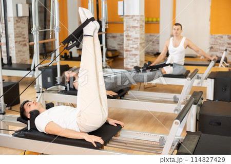 Man works out in gym and performs set of exercises to stretch leg muscles with help of reformer Man works out in gym and performs set of exercises to stretch leg muscles with help of reformer 114827429
