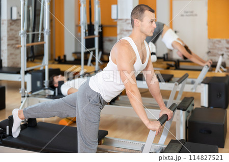 Man works out in gym and performs set of exercises to stretch leg muscles with help of reformer 114827521