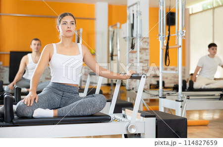 Girl sits on reformer and rests while doing exercises for muscles of body and torso. Girl sits on reformer and rests while doing exercises for muscles of body and torso. 114827659
