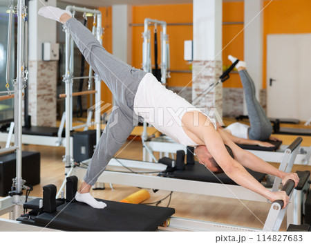 Man works out in gym and performs set of exercises to stretch leg muscles with help of reformer 114827683