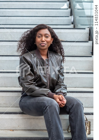 Woman in leather jacket sits comfortably on wooden stairs, smiling Woman in leather jacket sits comfortably on wooden stairs, smiling 114831044