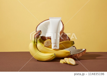 A unbranded tube of cosmetic displayed on a pile of fresh bananas and tropical coconut in the center of brown table over yellow background. Mockup scene for advertising with blank space for text A unbranded tube of cosmetic displayed on a pile of fresh bananas and tropical coconut in the center of brown table over yellow background. Mockup scene for advertising with blank space for text 114835196