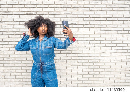 A woman in a denim jumpsuit taking a selfie against a brick wall backdrop A woman in a denim jumpsuit taking a selfie against a brick wall backdrop 114835994