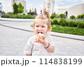 A hungry child enjoys eating ice cream outdoors. Portrait of a cute baby licking a creamy ice cream while walking in a city park, outdoor activities. 114838199