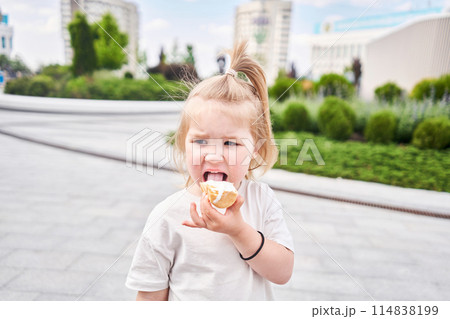 A hungry child enjoys eating ice cream outdoors. Portrait of a cute baby licking a creamy ice cream while walking in a city park, outdoor activities. A hungry child enjoys eating ice cream outdoors. Portrait of a cute baby licking a creamy ice cream while walking in a city park, outdoor activities. 114838199