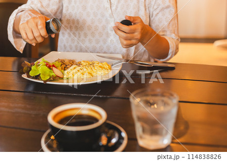 Woman having breakfast with hands putting black pepper on scrambled eggs. 114838826