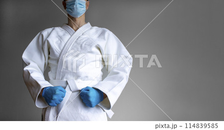COVID-19 Pandemic Coronavirus concept. A young strong man in a white kimono for sambo, jiu jitsu and other martial arts with a blue medical gloves and medical mask. Grey background. Copy space. 114839585